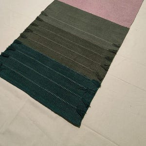 May include: A rectangular woven table runner featuring a color-blocked design with horizontal sections in deep teal, olive green, and soft pink. The fabric has a textured weave with small decorative fringe accents along the side edges of the runner.