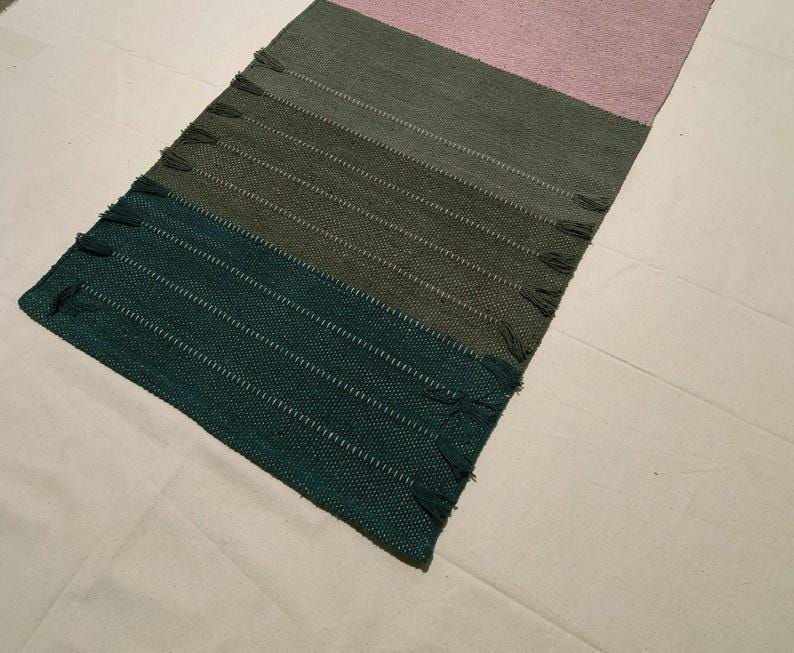 May include: A rectangular woven table runner featuring a color-blocked design with horizontal sections in deep teal, olive green, and soft pink. The fabric has a textured weave with small decorative fringe accents along the side edges of the runner.