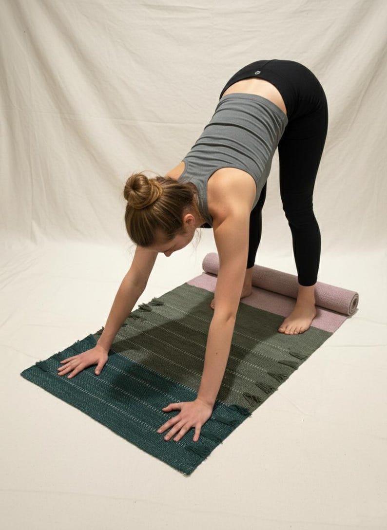 May include: A rectangular yoga mat featuring a color-blocked design with sections of dark teal, olive green, and dusty rose. The mat has a woven texture with small fringe details along the edges and is partially rolled at the top end.