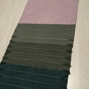May include: A rectangular woven table runner featuring horizontal color-blocked sections in shades of dark green, olive green, and a large central panel of soft dusty pink. The runner includes small decorative fringe tassels along the side edges.