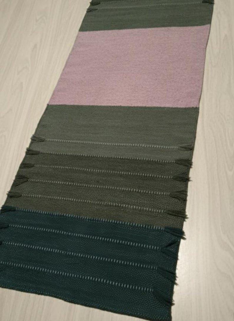 May include: A rectangular woven table runner featuring horizontal color-blocked sections in shades of dark green, olive green, and a large central panel of soft dusty pink. The runner includes small decorative fringe tassels along the side edges.