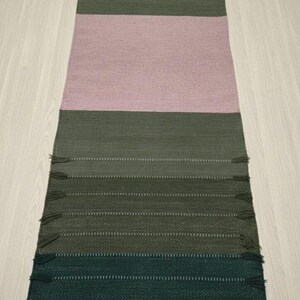 May include: A rectangular woven runner rug featuring horizontal color-blocked sections in shades of dark forest green, olive green, and a large central panel of soft dusty pink. The rug is finished with small, matching fringe tassels along the sides.