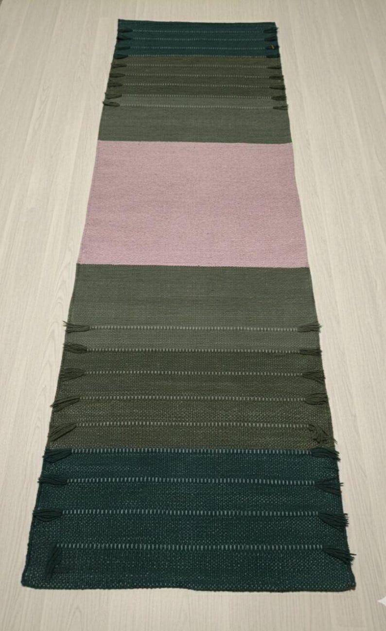 May include: A rectangular woven runner rug featuring horizontal color-blocked sections in shades of dark forest green, olive green, and a large central panel of soft dusty pink. The rug is finished with small, matching fringe tassels along the sides.