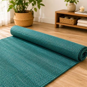 May include: A teal area rug with a woven texture, partially rolled up to reveal its construction. The rug features a consistent deep blue-green colour throughout its surface, showcasing a dense, braided pattern that creates a uniform appearance.