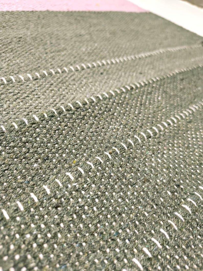 May include: A textured olive green woven fabric featuring horizontal rows of white stitched detailing. The material has a dense, nubby surface with a consistent pattern of parallel lines running across the length of the textile surface.