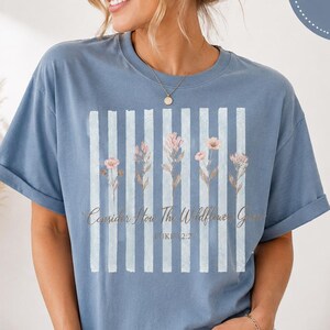Può includere: Una t-shirt blu a maniche corte con una grafica a righe verticali bianche e cinque delicati fiori di campo rosa. Il testo "Consider How The Wildflowers Grow" e "Luke 12:27" è stampato in corsivo al centro del disegno.
