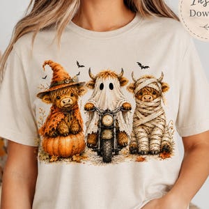 Può includere: Una t-shirt color crema con una grafica di tre mucche Highland in costume di Halloween: una strega su una zucca, un fantasma su una motocicletta e una mummia. Il testo "Instant Download" appare all'interno di un cerchio.