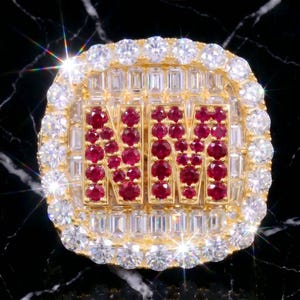 Könnte beinhalten: Ein goldener Ring mit den Buchstaben NM aus runden roten Steinen, umgeben von einer Einfassung aus klaren Steinen im Baguetteschliff und einem äußeren Rand aus runden klaren Steinen. Das Design liegt auf einem schwarzen Hintergrund mit weißer Marmorierung.