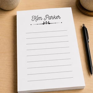 May include: A white rectangular notepad featuring the name Kim Parker printed in black script at the top, followed by a decorative horizontal line with a central emblem and eight evenly spaced horizontal lines for writing notes.
