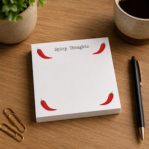 May include: A square white notepad featuring the text Spicy Thoughts printed in black at the top center. The notepad is decorated with four small red chili pepper illustrations, one positioned near each corner of the paper surface.
