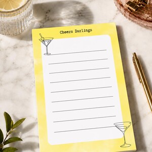 Può includere: Un blocco note rettangolare con bordo ad acquerello giallo e centro bianco con linee orizzontali. Il testo Cheers Darlings è stampato in alto. Illustrazioni a tratto nero di bicchieri da martini si trovano nell'angolo in alto a sinistra e in basso a destra.