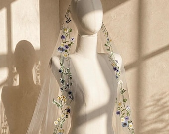 May include: A sheer, floor-length bridal veil draped over a mannequin, featuring delicate embroidery of climbing vines with small blue, purple, yellow, and white flowers along the edges of the lightweight, translucent tulle fabric.