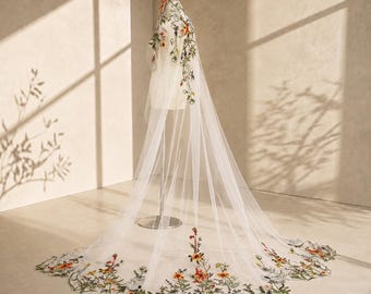 May include: A long, sheer white bridal veil draped over a mannequin. The veil features intricate, colorful floral embroidery in shades of orange, yellow, and green, concentrated at the top and along the scalloped bottom edge.