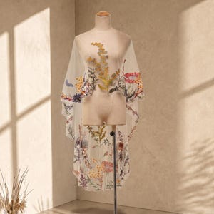Könnte beinhalten: Ein transparentes, weißes Capelet mit aufwendiger, mehrfarbiger Blumenstickerei. Das Design zeigt verschiedene Wildblumen in Gelb-, Rot-, Blau- und Grüntönen, die über den leichten, durchscheinenden Stoff mit einem gewellten Saum verteilt sind.