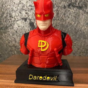 Puede incluir: Un busto impreso en 3D del personaje Daredevil, con un traje rojo con detalles en negro, un emblema amarillo en el pecho y ojos amarillos. La figura se asienta sobre una base rectangular negra con el texto Daredevil impreso en amarillo.