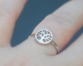 May include: A silver ring featuring a circular tree of life design. The ring's band is thin and delicate, and the tree design is open, allowing light to pass through. The ring is worn on a finger with red nail polish.