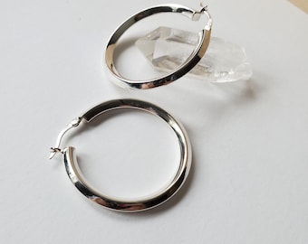 May include: Two silver hoop earrings with a polished finish. The earrings are round and have a simple, classic design. They are displayed on a white surface with a clear crystal in the background. Jewelry.