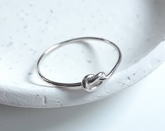 May include: A silver knot ring with a simple design. The ring is thin and delicate, and the knot is smooth and polished.