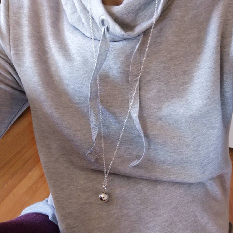 May include: A silver chain necklace with a round silver pendant. The necklace is worn over a grey hooded sweatshirt.