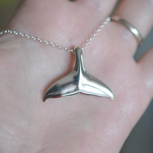 May include: A silver-colored necklace featuring a whale tail pendant. The pendant is smooth and polished, reflecting light. The chain is delicate and links to the tail. The necklace is shown against a light background.
