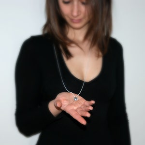 May include: A woman wearing a black top with a silver chain necklace and a round pendant. She is holding the pendant in her hand.