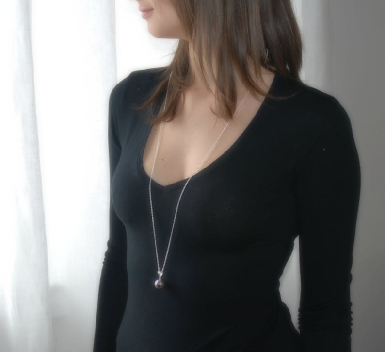 May include: A silver chain necklace with a round silver pendant. The necklace is worn by a person with long brown hair wearing a black v-neck top.