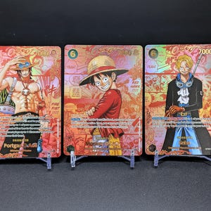 May include: Three One Piece trading cards featuring Portgas D. Ace, Monkey D. Luffy, and Sabo. Each card has a gold-toned holographic finish with manga-style panels, character names, power levels of 7000, and text in English on a red background.