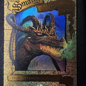 Może przedstawiać: Prostokątna karta kolekcjonerska ze złotą obwódką i napisem Smaug the Magnificent. Centralna ilustracja przedstawia brązowego smoka z czerwonymi oczami i zielonym dymem wydobywającym się z pyska. Dolny tekst brzmi LEGENDARY CREATURE - DRAGON.