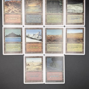 May include: A collection of ten rectangular trading cards arranged in three rows. Each card features a unique landscape illustration, a white border, and a text box containing specific game rules and coloured geometric symbols at the bottom.
