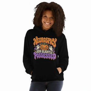 May include: A black hooded sweatshirt featuring a graphic print with the text "NEUROSPICY AND SLIGHTLY POSSESSED" in orange and purple. The design includes a central jack-o'-lantern pumpkin, two small white ghosts, and small yellow stars.