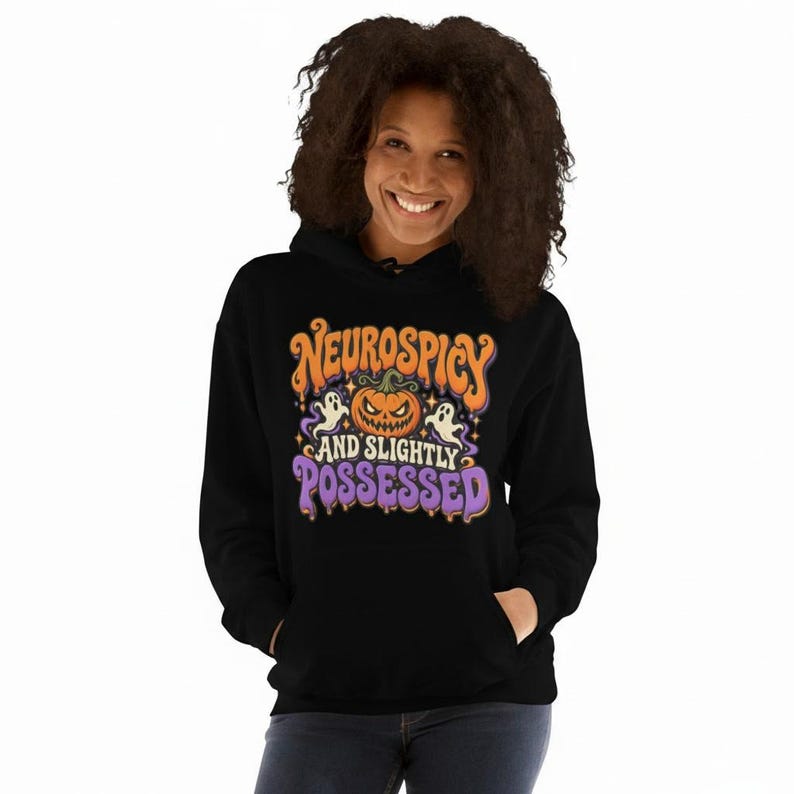 May include: A black hooded sweatshirt featuring a graphic print with the text "NEUROSPICY AND SLIGHTLY POSSESSED" in orange and purple. The design includes a central jack-o'-lantern pumpkin, two small white ghosts, and small yellow stars.