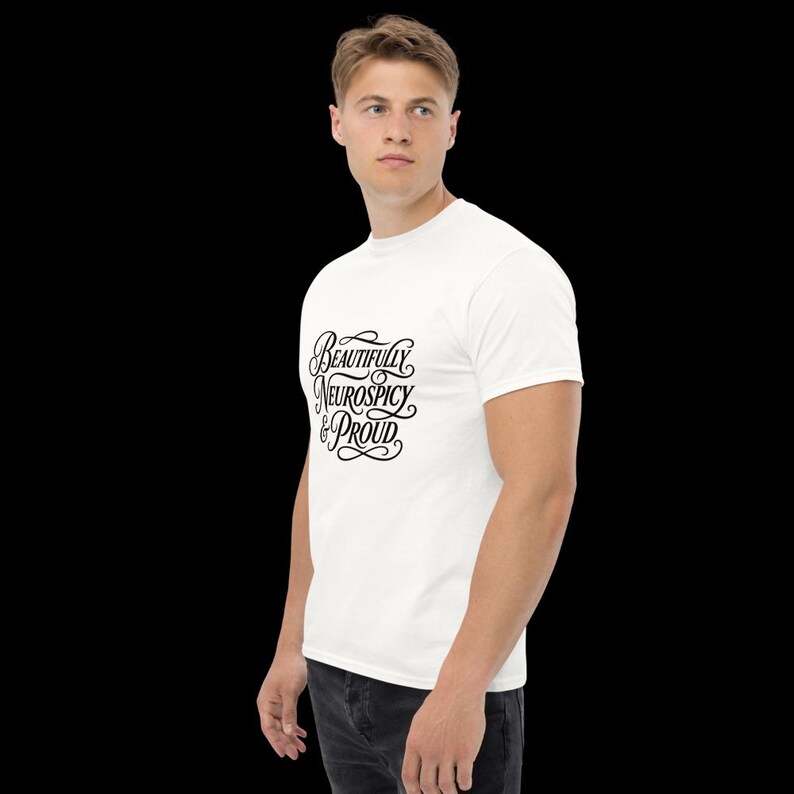 K&ouml;nnte beinhalten: Ein wei&szlig;es kurz&auml;rmeliges T-Shirt mit schwarzem, kursivem Text auf der Brust, der Beautifully Neurospicy & Proud lautet. Das T-Shirt hat einen Rundhalsausschnitt und eine normale Passform, kombiniert mit einer dunkelgrauen Jeans.