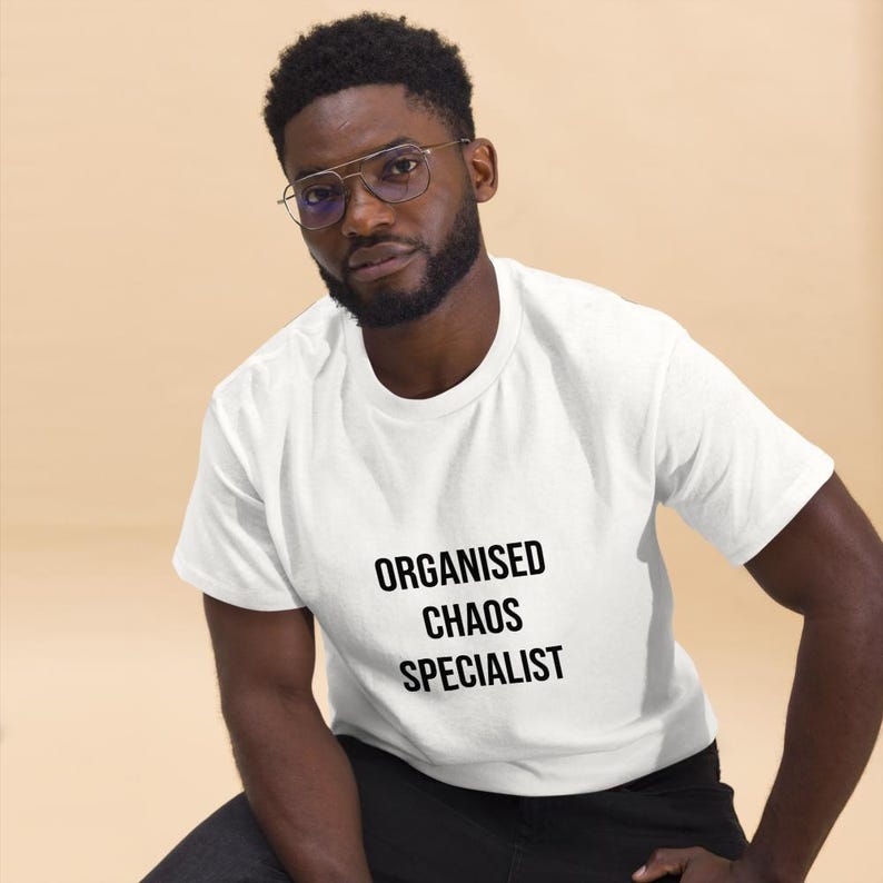 May include: A white short-sleeved t-shirt featuring the text "ORGANISED CHAOS SPECIALIST" printed in bold black capital letters on the front. The shirt has a classic crew neckline and a standard fit, providing a clean and simple appearance.