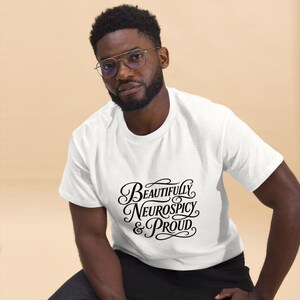 K&ouml;nnte beinhalten: Ein wei&szlig;es Kurzarm-T-Shirt mit schwarzer Schreibschrift auf der Brust, die Beautifully Neurospicy & Proud lautet. Der Text ist in drei Zeilen angeordnet und mit dekorativen Schn&ouml;rkeln an den Wortanf&auml;ngen und -enden versehen.