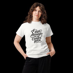 Może przedstawiać: Biały t-shirt z krótkim rękawem z czarnym napisem kursywą na klatce piersiowej o treści Error messages are my comfort genre. Koszulka ma okrągły dekolt i jest zestawiona z gładkimi czarnymi spodniami, tworząc prostą i przejrzystą estetykę.