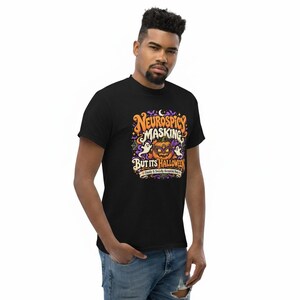 Pu&ograve; includere: Una t-shirt nera con una grafica colorata che riporta il testo "NEUROSPICY MASKING BUT ITS HALLOWEEN" e "A FINALLY A SOCIALLY ACCEPTABLE MASK". Il disegno include una zucca con una maschera, fantasmi e stelle.