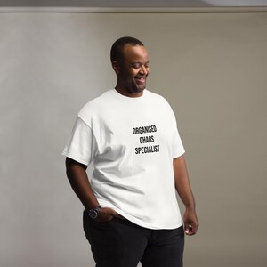 May include: A white short-sleeved t-shirt featuring the black text "ORGANISED CHAOS SPECIALIST" printed on the chest. The garment has a crew neckline and a standard fit, paired with dark trousers and a wristwatch on the left wrist.