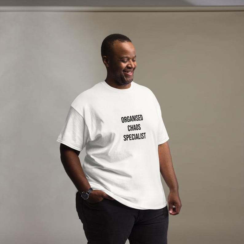 May include: A white short-sleeved t-shirt featuring the black text "ORGANISED CHAOS SPECIALIST" printed on the chest. The garment has a crew neckline and a standard fit, paired with dark trousers and a wristwatch on the left wrist.
