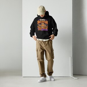 May include: A black hooded sweatshirt featuring a graphic print with the text Happy Halloween You Neurospicy Little Gremlins in orange, purple, and white. The hoodie is paired with khaki cargo trousers, a beige baseball cap, and white trainers.