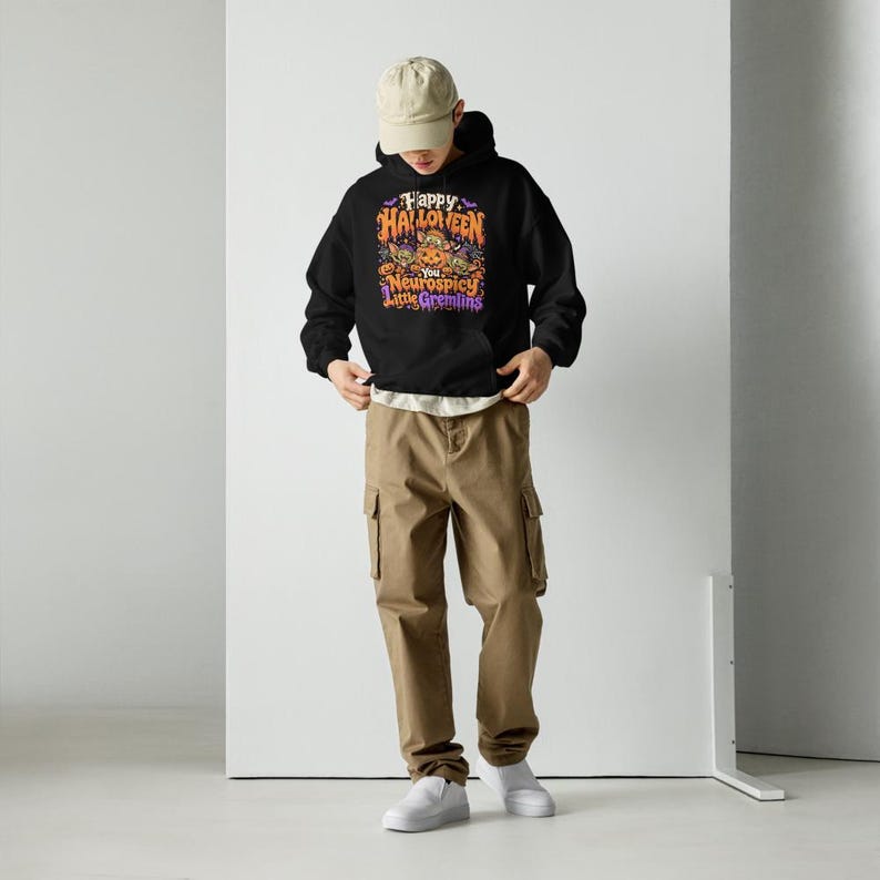 May include: A black hooded sweatshirt featuring a graphic print with the text Happy Halloween You Neurospicy Little Gremlins in orange, purple, and white. The hoodie is paired with khaki cargo trousers, a beige baseball cap, and white trainers.