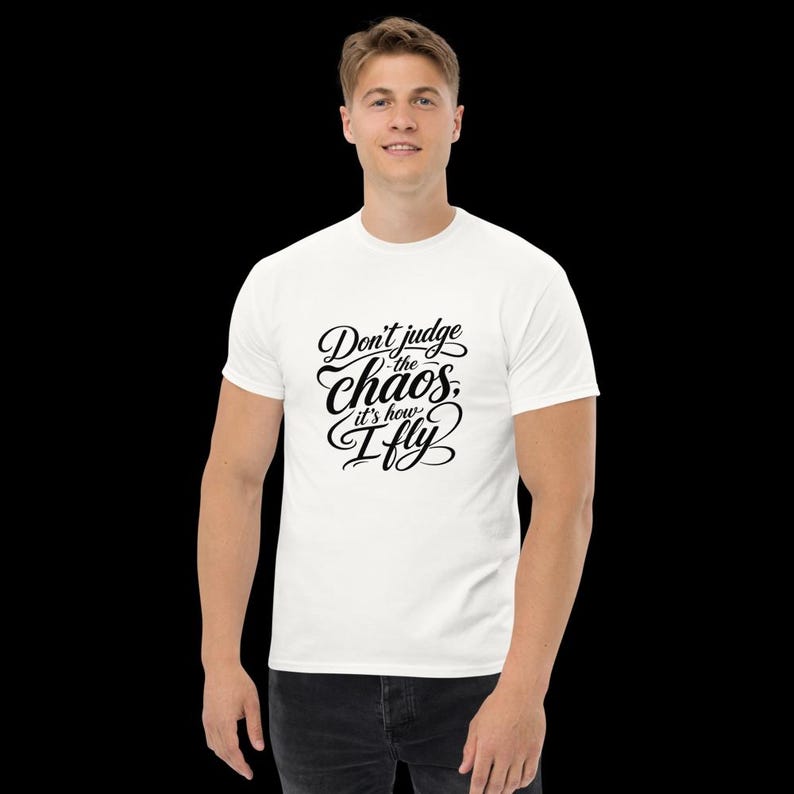 May include: A white short-sleeved t-shirt featuring black cursive text on the chest that reads, Don't judge the chaos, it's how I fly. The shirt has a crew neckline and a standard fit, providing a clean look with bold, stylised lettering.