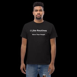 K&ouml;nnte beinhalten: Ein schwarzes Kurzarm-T-Shirt mit dem wei&szlig;en Text "I Like Routines" &uuml;ber einer horizontalen Linie und dem Text "More Than People" direkt darunter. Das T-Shirt wird mit einer blauen Jeans kombiniert, die ein abgenutztes Detail aufweist.