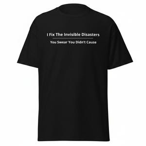 Op de afbeelding: Een zwart t-shirt met korte mouwen en witte tekst op de borst met de tekst: I Fix The Invisible Disasters, met een horizontale lijn eronder, gevolgd door de tekst: You Swear You Didn't Cause, in een strak schreefloos lettertype.
