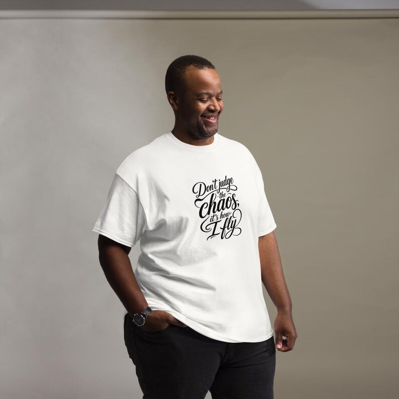 May include: A white short-sleeved t-shirt featuring black cursive and print text on the chest that reads, Don't judge the chaos, it's how I fly. The shirt has a crew neckline and a standard fit, providing a clean and simple look for everyday wear.