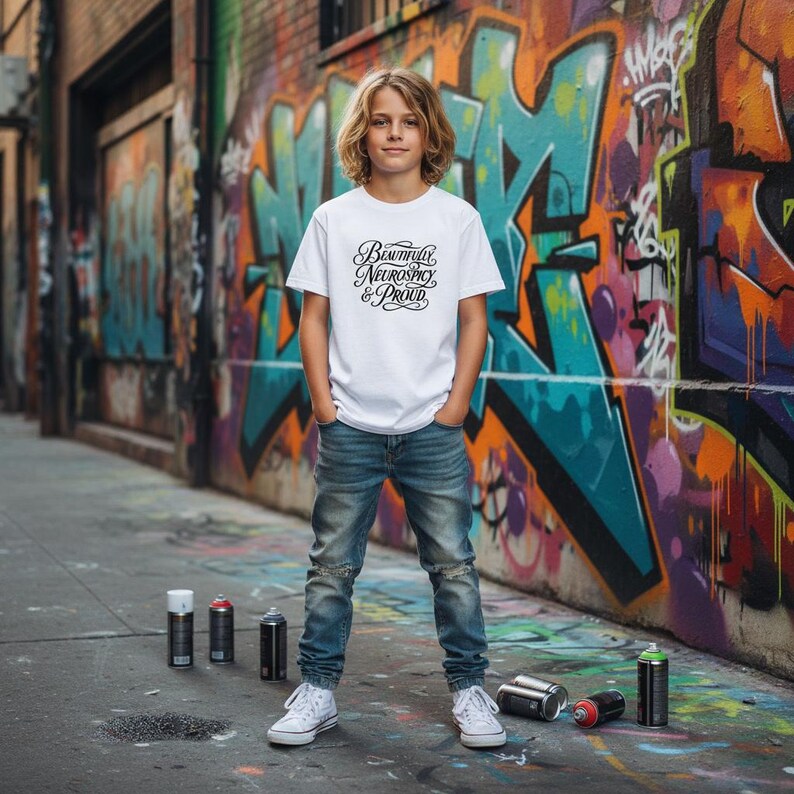 K&ouml;nnte beinhalten: Ein wei&szlig;es kurz&auml;rmeliges T-Shirt mit dem schwarzen Schriftzug "Beautifully Neurospicy & Proud" in einer stilisierten Schreibschrift, kombiniert mit einer blauen Jeans und wei&szlig;en Turnschuhen, in einer st&auml;dtischen Umgebung mit Spr&uuml;hdosen.