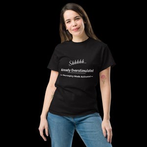 May include: A black short-sleeved t-shirt featuring white text that reads Shhhhh... Already Overstimulated - Neurospicy Mode Activated - centred on the chest. The shirt is paired with blue denim jeans, creating a casual and simple outfit.