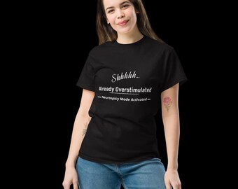May include: A black short-sleeved t-shirt featuring white text that reads Shhhhh... Already Overstimulated - Neurospicy Mode Activated - centred on the chest. The shirt is paired with blue denim jeans, creating a casual and simple outfit.
