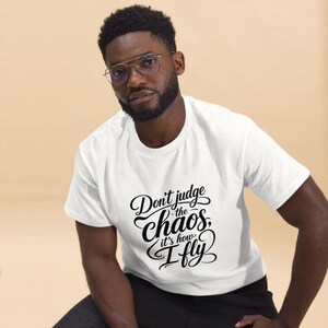 May include: A white short-sleeved t-shirt featuring black cursive and bold lettering on the chest that reads, Don't judge the chaos, it's how I fly. The shirt has a classic crew neckline and a standard fit, providing a clean and simple look.