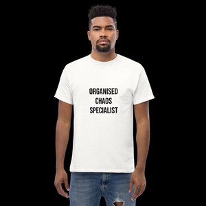 May include: A white short-sleeved t-shirt featuring the black text "ORGANISED CHAOS SPECIALIST" printed in a clean, sans-serif font on the chest. The garment is paired with blue denim jeans that have a visible distressed detail at the knee.
