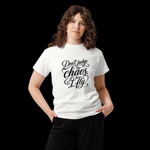 May include: A white short-sleeved t-shirt featuring black cursive text on the front that reads, Don't judge the chaos, it's how I fly. The shirt has a crew neckline and a standard fit, paired with black trousers.